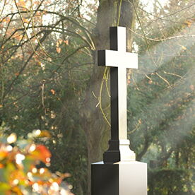 Sonnenstrahlen fallen auf einen mit Bäumen umringten Grabstein in Form eines Kreuzes