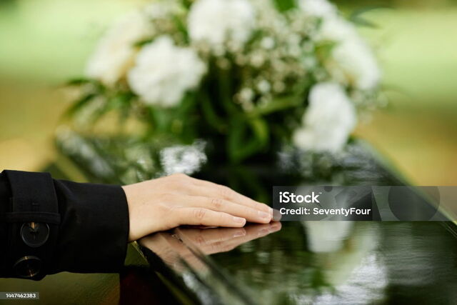 Hand streicht über einen schwarzen Sarg, im Hintergrund sind weiße Blumen auf dem Sarg liegend zu sehen